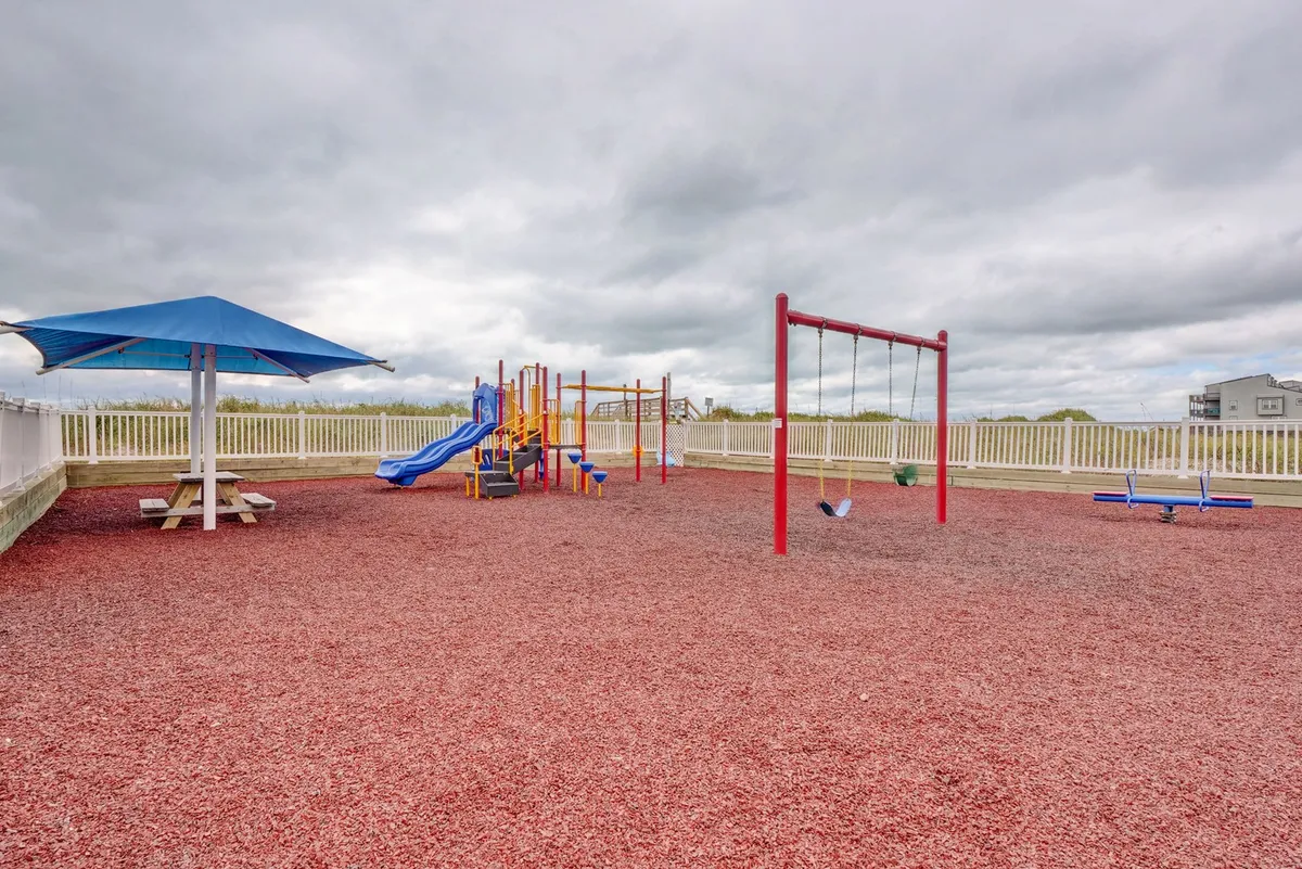 A colorful playground with swings and picnic area, surrounded by a safety fence. The scene is lively with opportunities for children to play and for families to gather outdoors.
