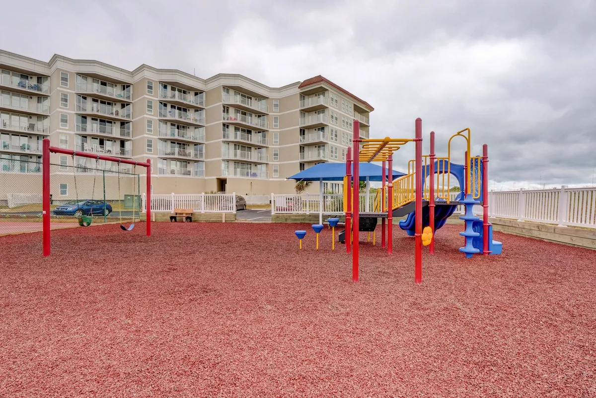 A vibrant, colorful playground with swings and modern play equipment sits on a soft, red rubber ground cover. Kids are shown playing, with residential buildings in the background, creating a joyful, community-oriented atmosphere.