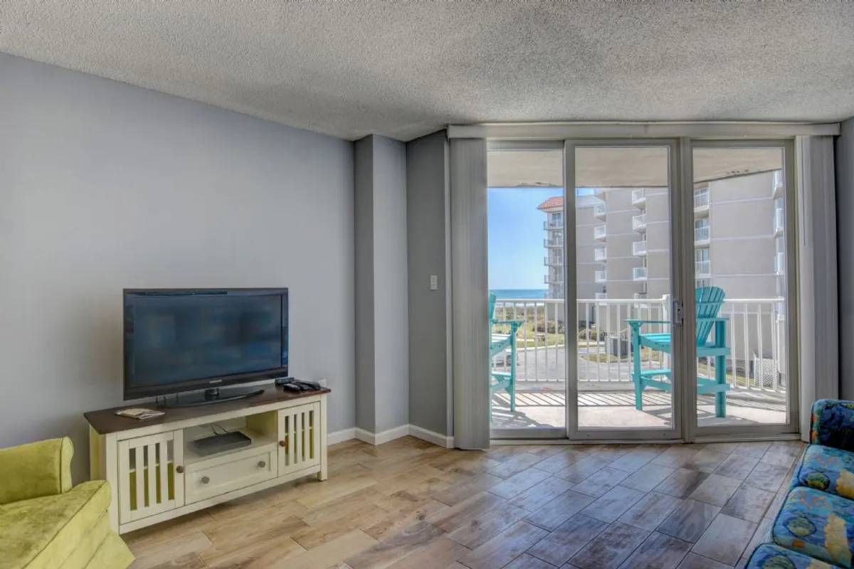 A contemporary interior scene highlighting a stylish television area, sliding glass doors leading out to a balcony, and blue chairs positioned on the balcony for enjoying views. The layout naturally draws the eye outdoors, reinforcing the connection between indoor comfort and outdoor serenity.