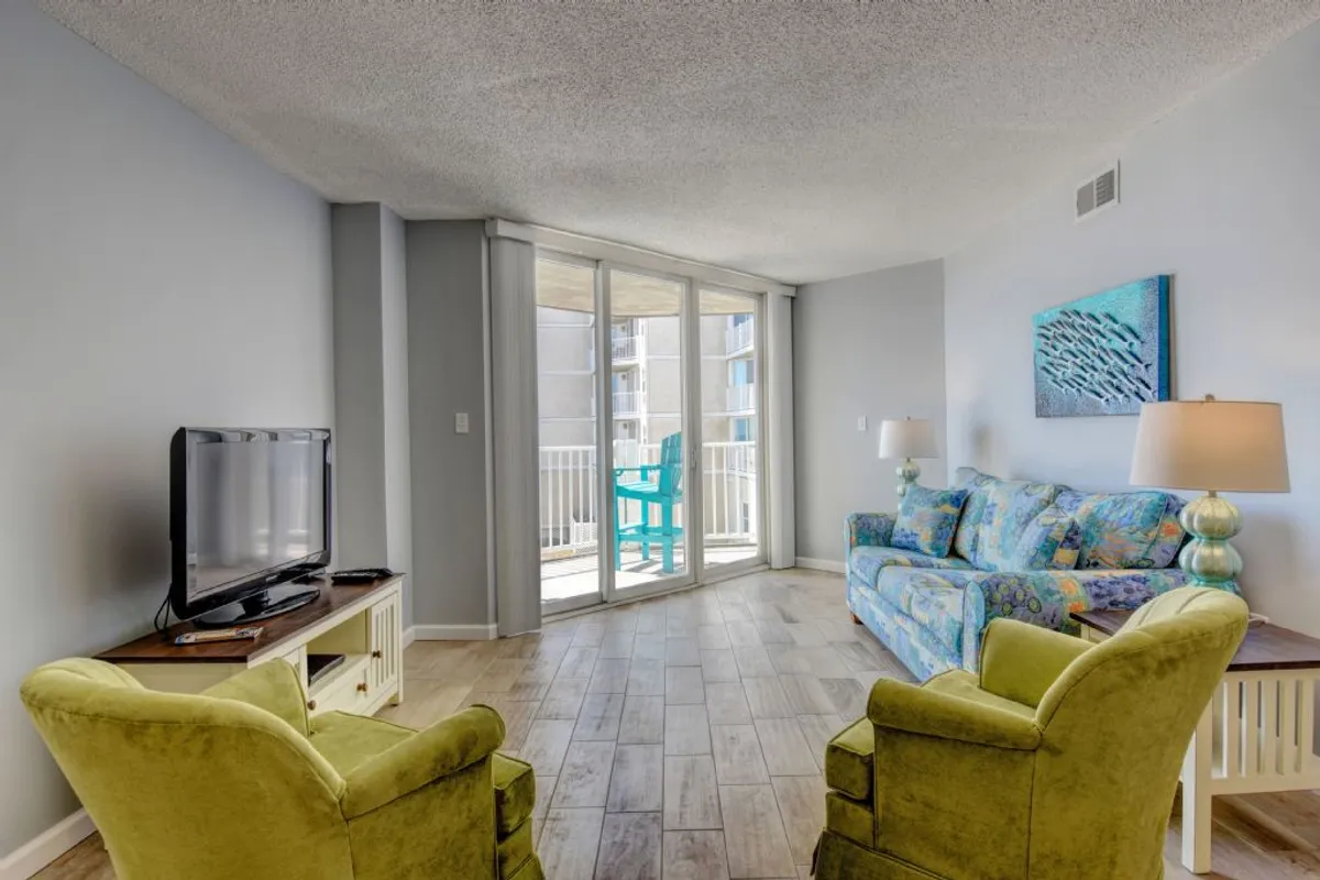 A casual and comfortable living room featuring a plush sofa, green accent chairs, a television ideal for movie nights, and expansive balcony doors that reveal a sweeping view outside. Natural light floods the space, and the open layout encourages relaxation, conversation, and easy transitions to the outdoors.