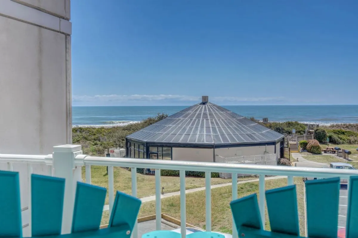 The image showcases a stunning ocean view from a private condo balcony, with comfortable outdoor furniture inviting guests to relax. A unique round building anchors the scene, surrounded by lush green lawns, all set against the backdrop of the sparkling Atlantic on North Topsail Beach. The tranquil setting hints at moments of sunrise yoga, sunset gatherings with friends, and peaceful reflection by the sea.