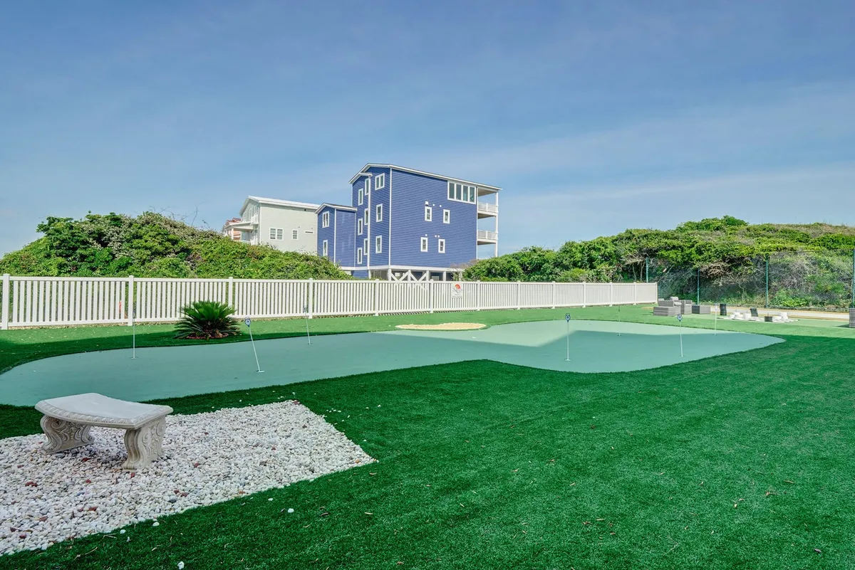 The image showcases an artificial putting green surrounded by manicured landscaping and a stone bench, with a blue house in the background. The atmosphere is both playful and relaxed, suggesting opportunities for golf practice or leisurely social moments outdoors.