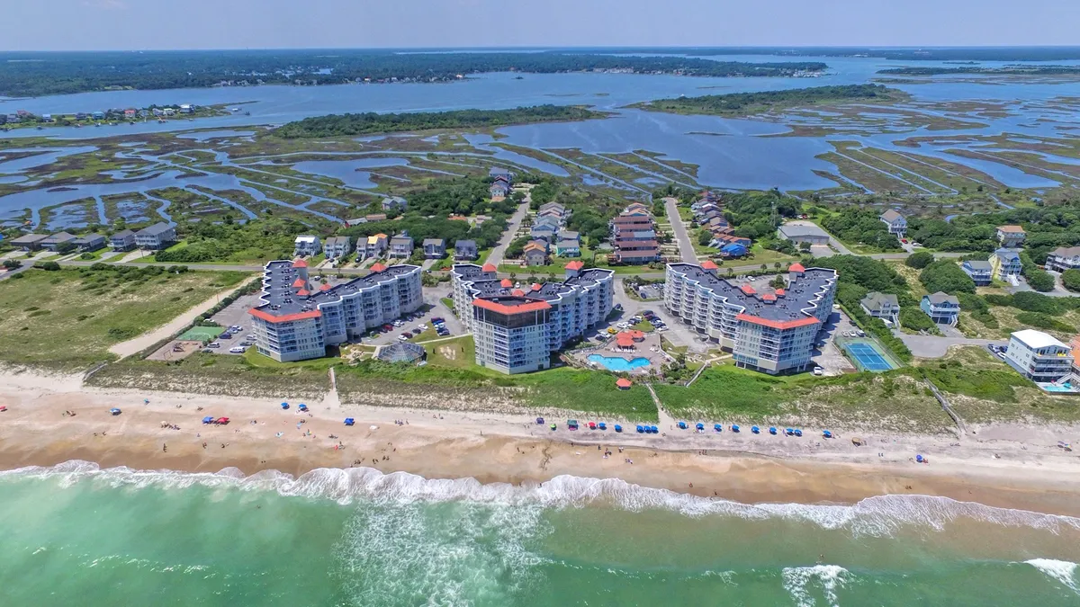 This image showcases the resort's beachfront condominiums with a sparkling swimming pool in the foreground, shaded beach umbrellas along the sand, and views extending to marshland and open coastline. The scene captures guests enjoying poolside relaxation, sunbathing, and leisurely strolls or gatherings by the beach as the sun sets.