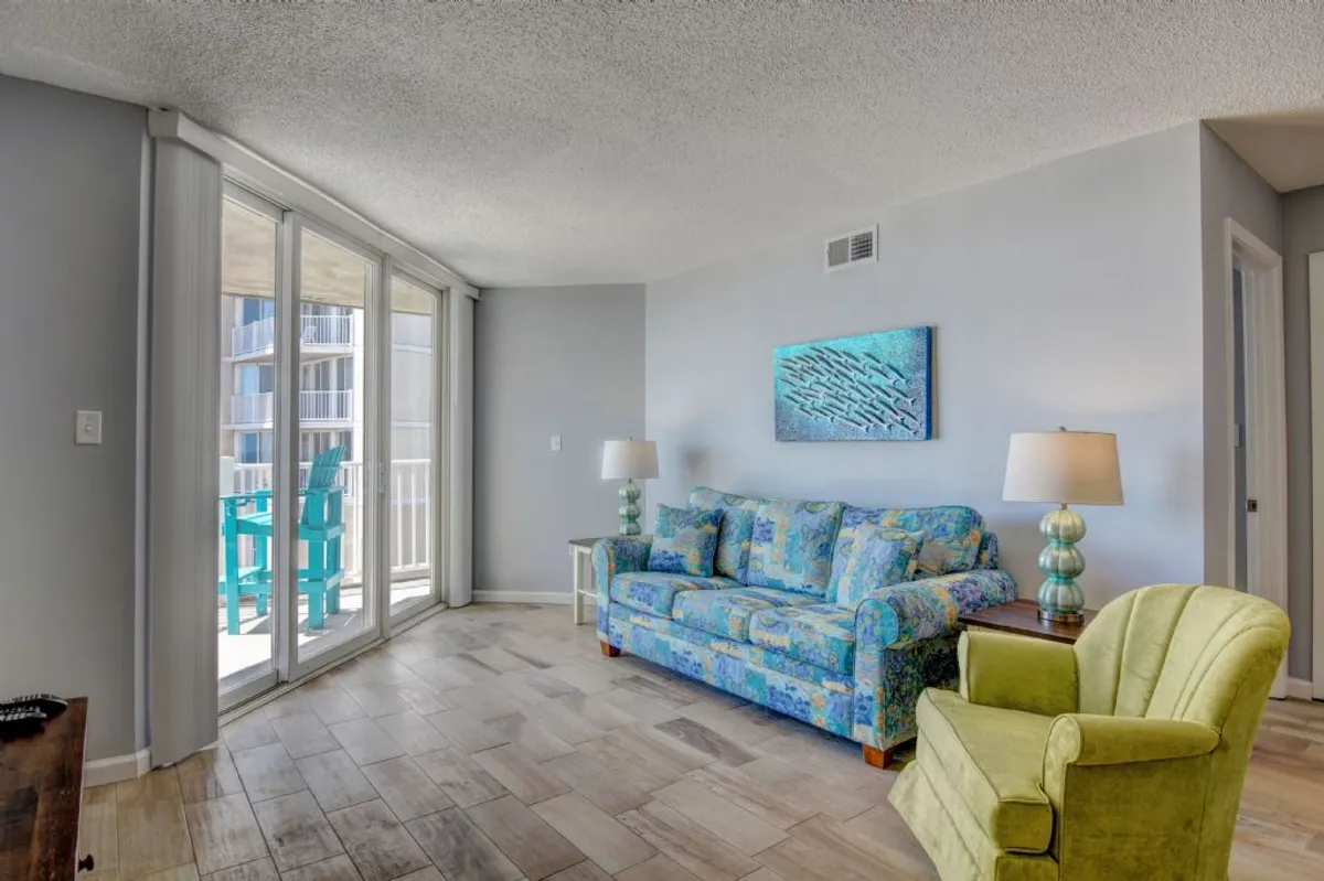 A cheerful, welcoming living area with a colorful couch, green armchair, large windows, and abstract wall art. The room exudes warmth and relaxation, with ample natural light and a direct sightline to the outdoors, hinting at an airy, social atmosphere.