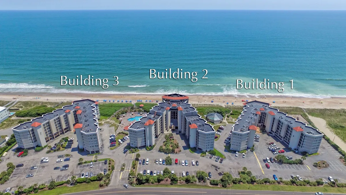 An exterior view featuring three resort buildings, a large outdoor swimming pool, and a sweeping ocean backdrop. The resort amenities, including the pool and proximity to the beach, are visible, all framed in a vacation and leisure atmosphere ideal for families.