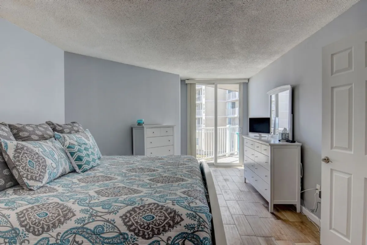A serene bedroom featuring a bed with patterned bedding, large windows that let in natural light, and a dresser with a mirror. The tranquil ambiance is enhanced by soft, neutral décor, inviting relaxation and restful sleep.